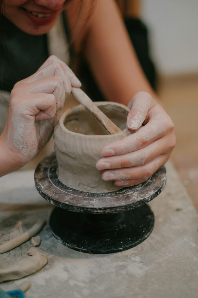 Een vrolijke vrouw werkt aan een handvorm object van klei. Het object staat op een draai plateau en ze houd het vast met haar hand. Met haar andere hand bewerkt ze de klei met een houten mes