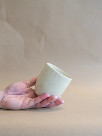 Hand houdt handgemaakte mok in Beige glazuur van Iris Floor Ceramics schuin in de lucht, met een warme naturel tint die de organische schoonheid van de klei benadrukt.