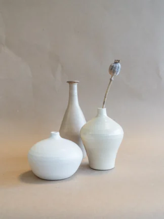 Drie handgemaakte vazen in Beige, Matte White en Velvet Beige van Iris Floor Ceramics, met de Matte White ballonvormige vaas vastgehouden door een hand, met de onderkant zichtbaar, tegen een naturel beige bruine achtergrond.