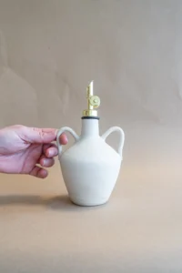 Het olie flesje in Matte White staat op tafel en wordt vastgehouden aan een oortje. Het flesje heeft een kruikvorm en twee oortjes, handgemaakt door Iris Floor Ceramics.