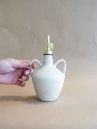 Het olie flesje in Matte White staat op tafel en wordt vastgehouden aan een oortje. Het flesje heeft een kruikvorm en twee oortjes, handgemaakt door Iris Floor Ceramics.