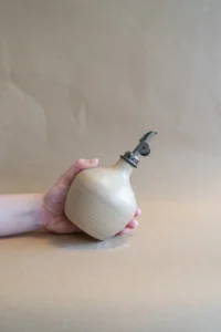 Hand houdt olie flesje in Velvet Beige glazuur schuin vast, alsof het wordt gebruikt om te schenken, met een schenk tuit in Gun Metal zwart, tegen een beige achtergrond.